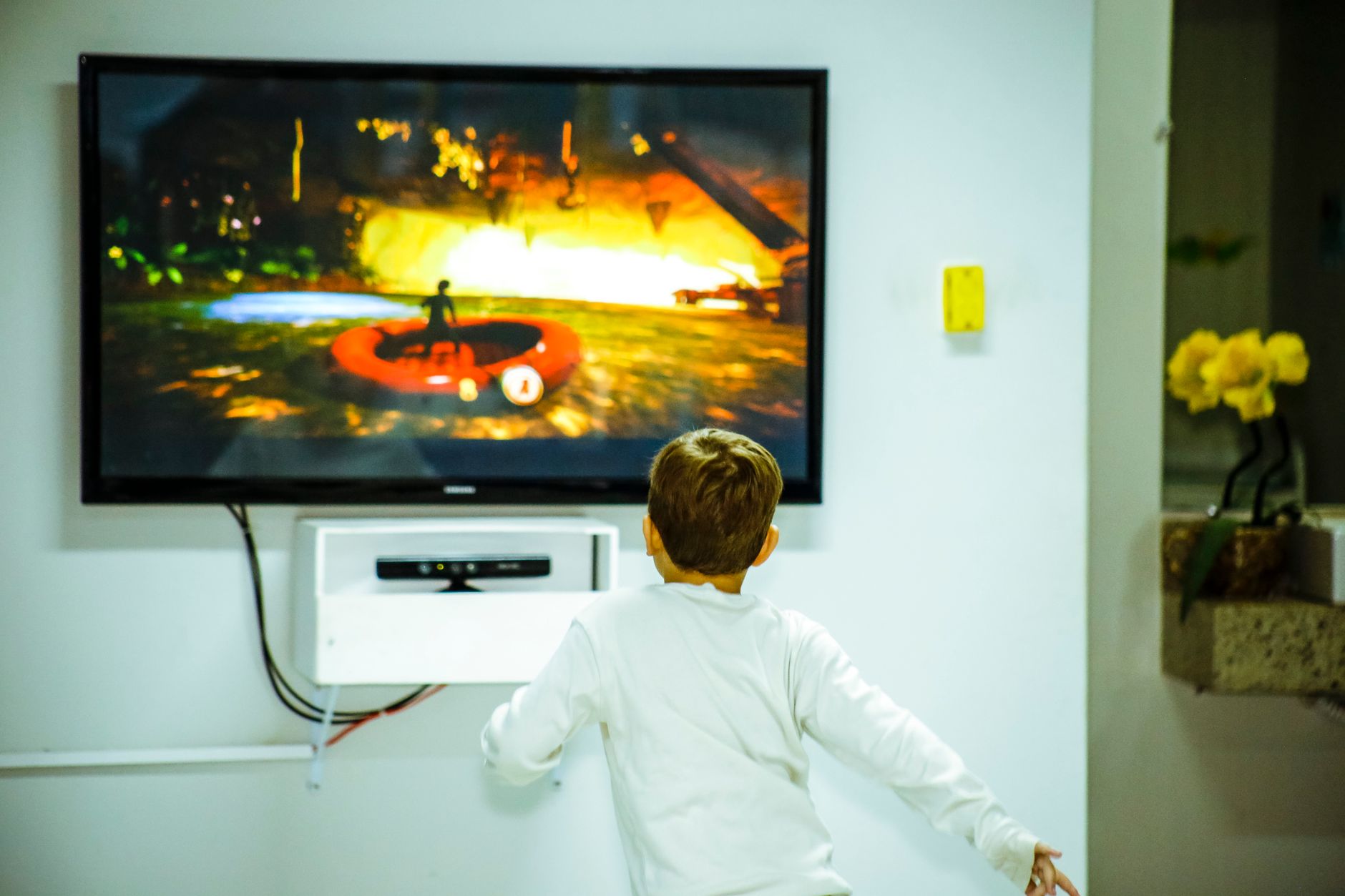 A young child in a white shirt is playing a video game, watching a television screen featuring a character in a red inflatable raft on a colorful landscape.