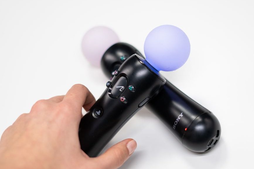 Close-up of a hand holding a Sony motion controller, with another controller in the background, both featuring colored light endpoints.