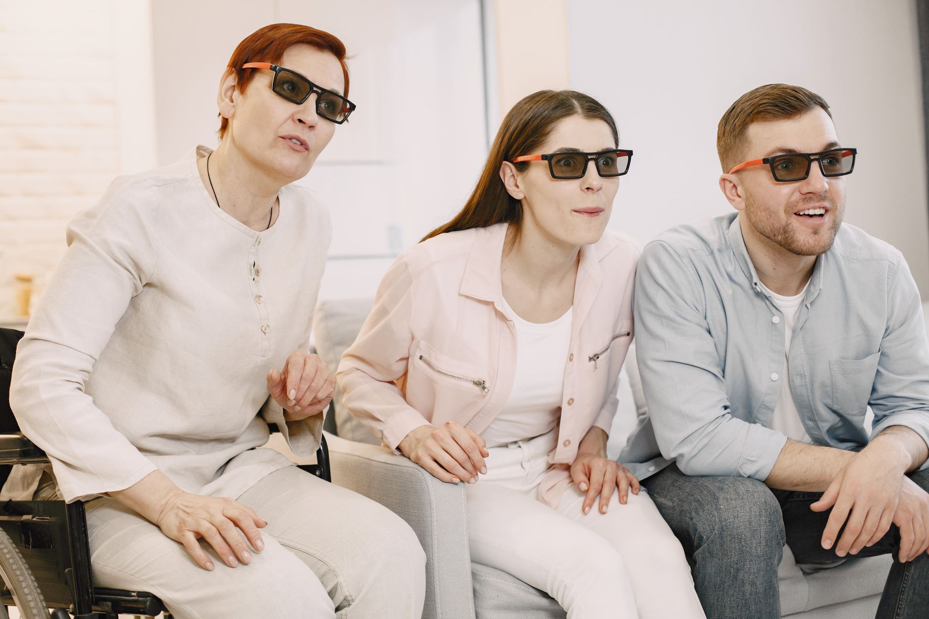 Three people watching a movie with 3D glasses, sitting closely together in a cozy living room. One person is in a wheelchair, while the others are seated and focused on the screen.
